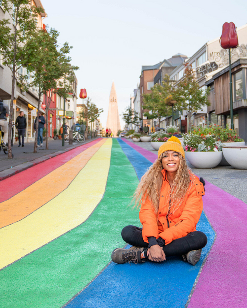 A person in a bright orange jacket sits on a road painted in rainbow colors.